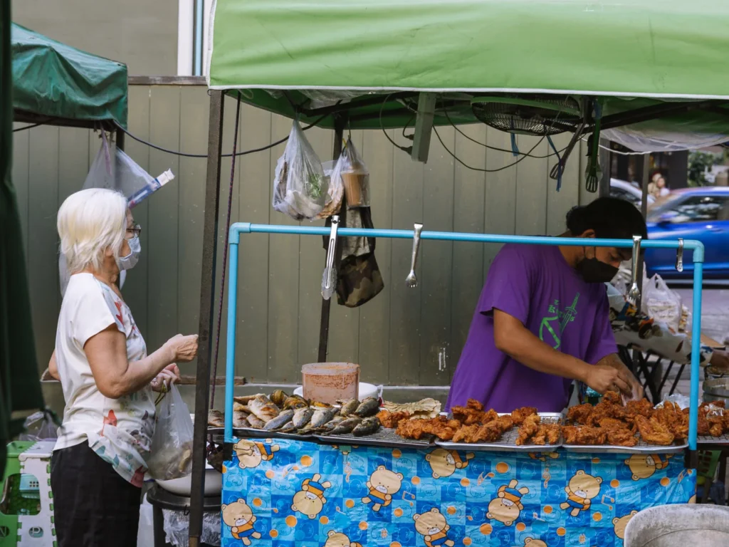 Woman buys fried seafood from masked vendor at Bangkok street food stall under green canopy.