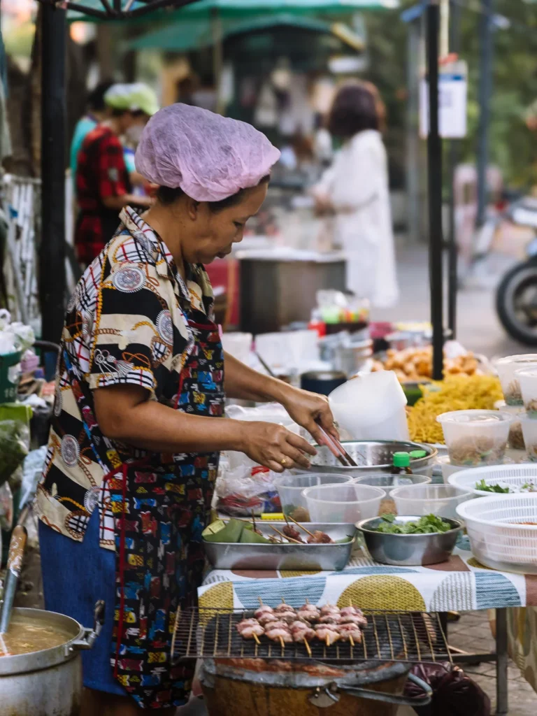 Vendor in colorful apron cooks noodles and skewers at vibrant Bangkok street food stall.