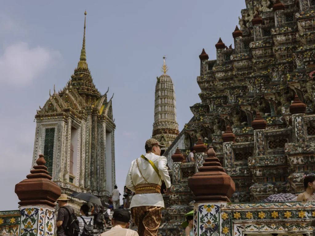 Visitor in Thai attire ascends Wat Arun's mosaic steps in Bangkok under a clear sky.
