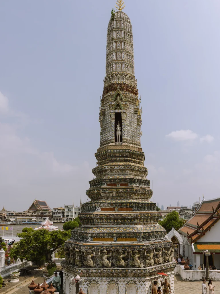 Close-up of Wat Arun's central prang tower in Bangkok, featuring intricate porcelain mosaics, guardian statues, and lush greenery under a blue sky.