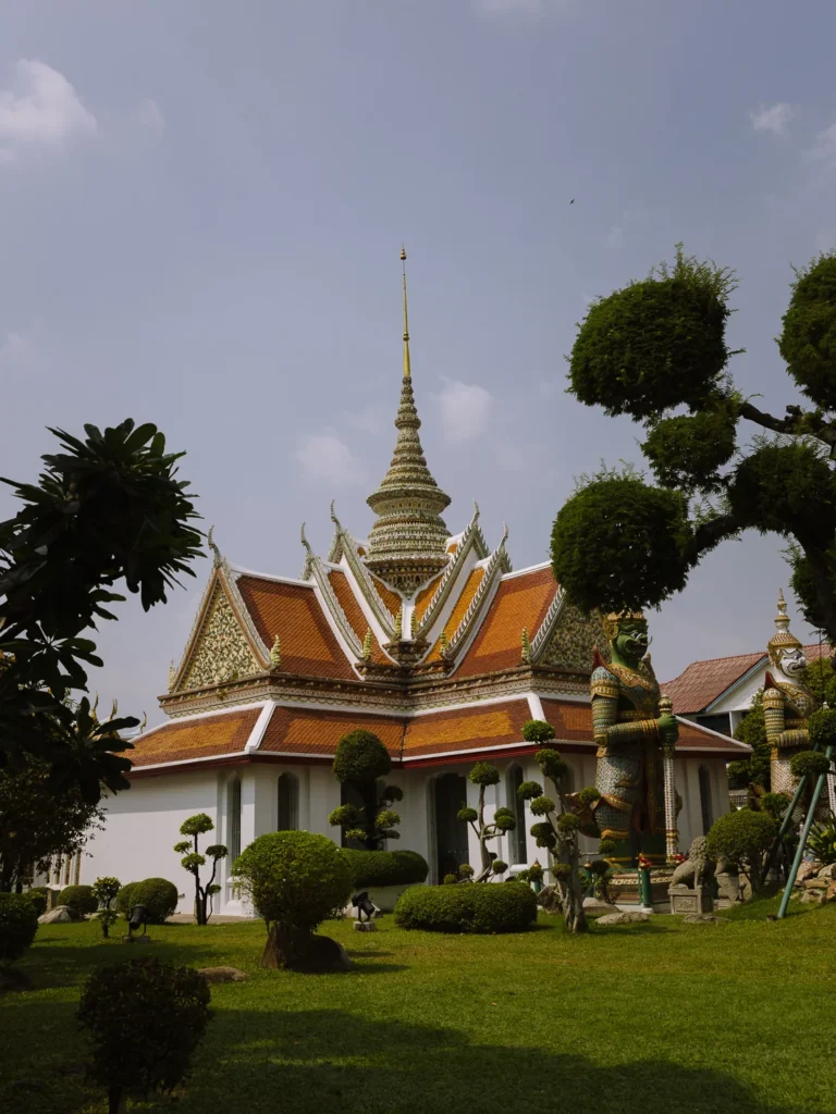 Ornate Thai pavilion at Wat Arun in Bangkok, with golden roof, guardian statues, and manicured bushes on green lawn under blue sky.