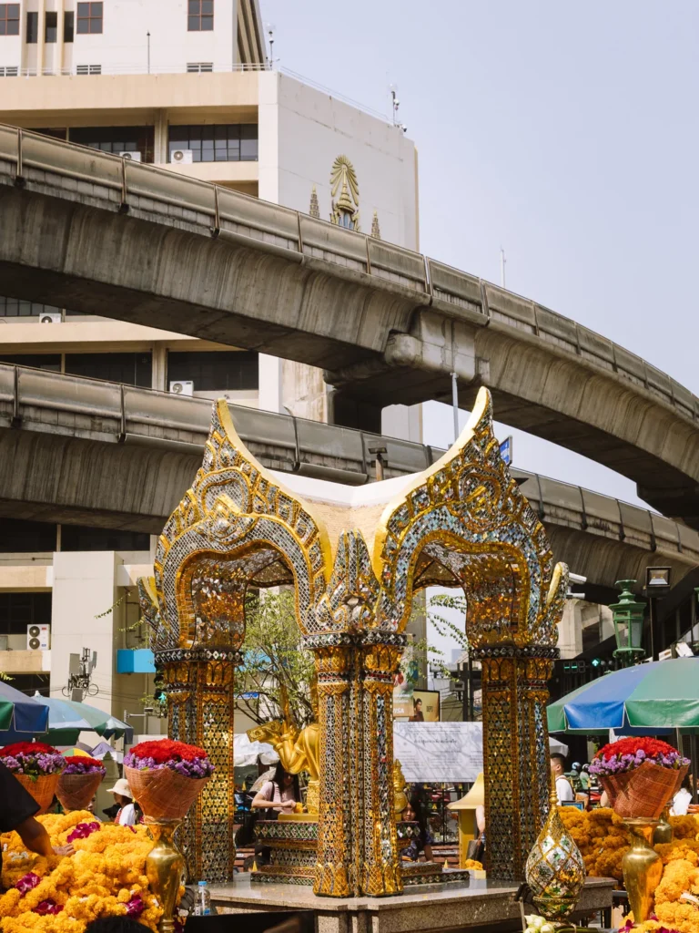 The ornate gold mosaic structure of the Erawan Shrine surrounded by marigold garlands, with a BTS elevated rail track curving overhead.