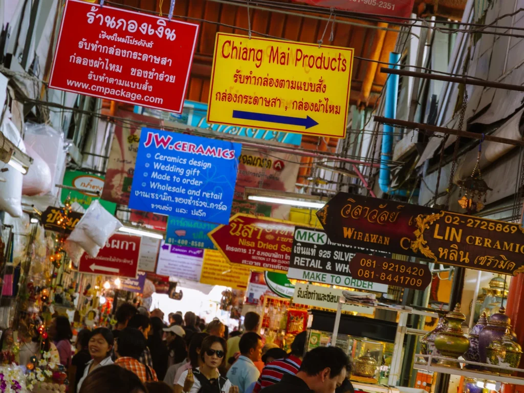 Crowds browse a busy covered aisle at Chatuchak Weekend Market, Bangkok, beneath a tangle of colourful vendor signs in Thai and English.