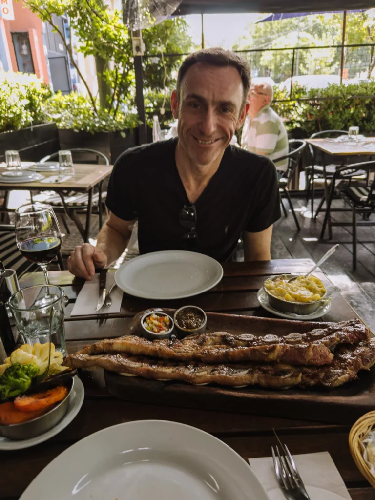 Mark smiling at an outdoor parrilla in Buenos Aires, with a large tira de asado (short ribs) on a wooden board in front of him, accompanied by a glass of red wine and side dishes