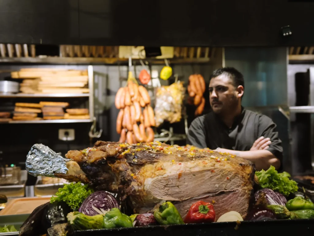 A Buenos Aires parrilla chef standing behind an enormous slow-roasted joint of beef on a grill, surrounded by vegetables, with strings of chorizos hanging in the kitchen behind him