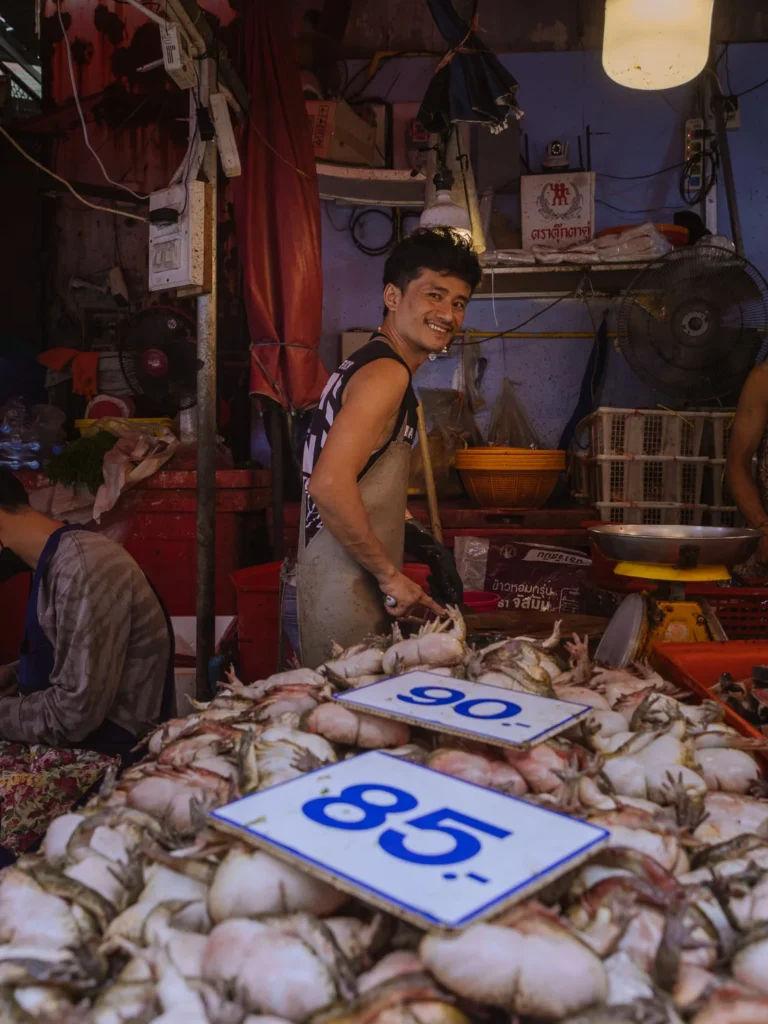 A smiling vendor stands behind a large pile of squid at a seafood stall in Khlong Toei Market, Bangkok.
