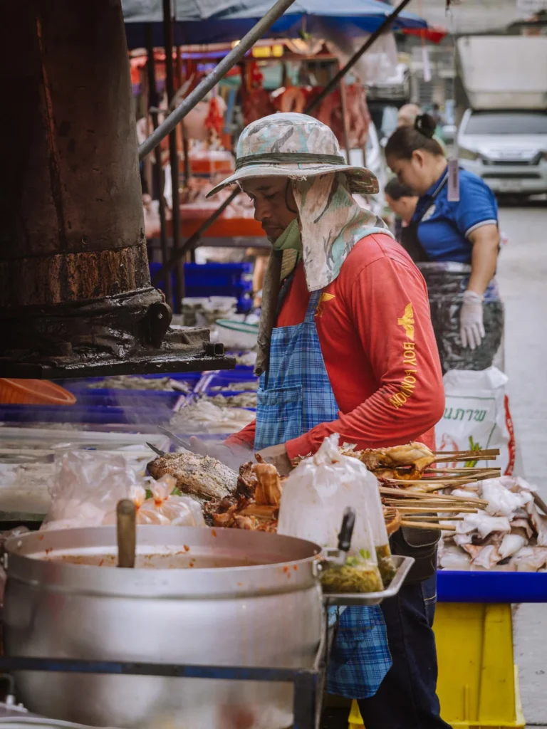A vendor prepares grilled fish and skewers at an outdoor stall in Khlong Toei Market, Bangkok.