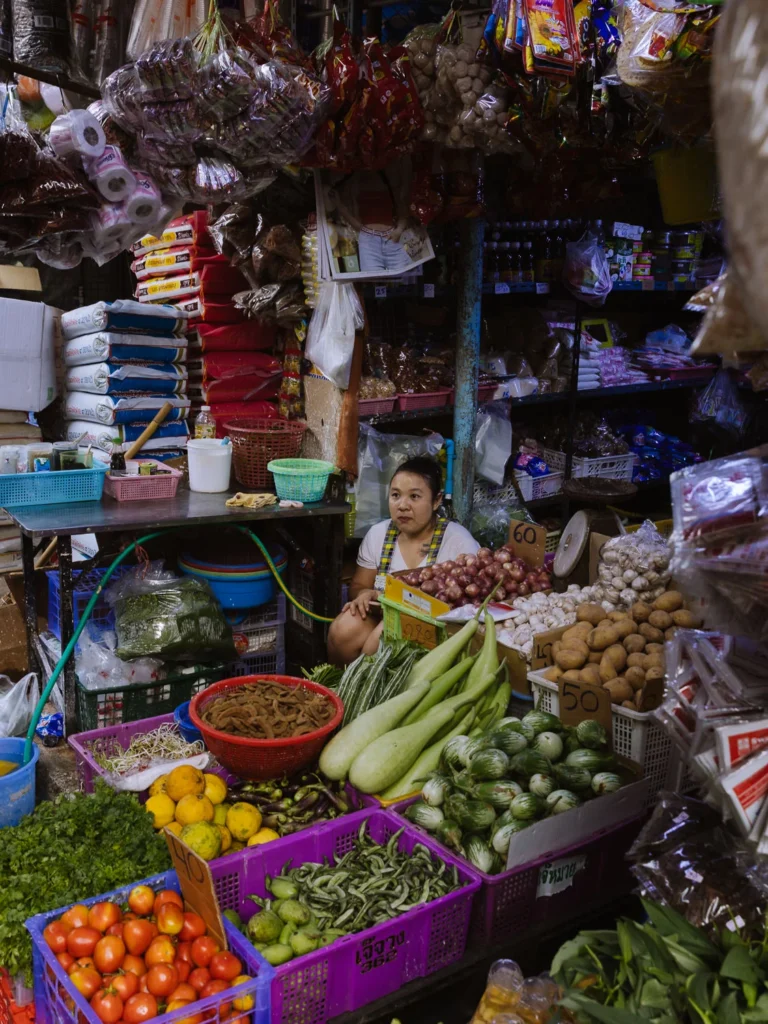 A vendor sits surrounded by fresh vegetables, dried goods, and produce piled high at a stall in Khlong Toei Market, Bangkok.