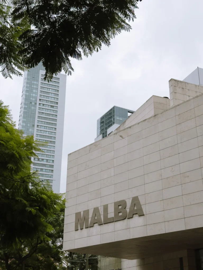 The exterior facade of MALBA (Museo de Arte Latinoamericano de Buenos Aires) in Palermo, with the museum's name in large letters on the stone cladding and glass towers visible behind