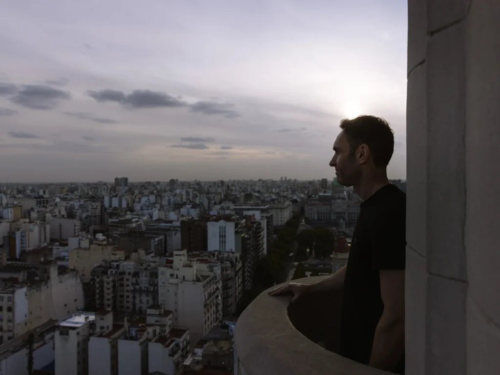 Mark looking out over the Buenos Aires cityscape from the observation balcony of the Palacio Barolo on Avenida de Mayo, silhouetted against a hazy dusk sky with the city's vast low-rise skyline stretching to the horizon