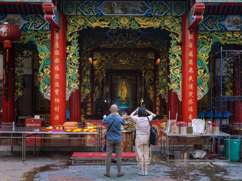 Two visitors photograph a golden deity statue inside a ornate Chinese shrine in Bangkok's Chinatown, framed by red columns with gold calligraphy.