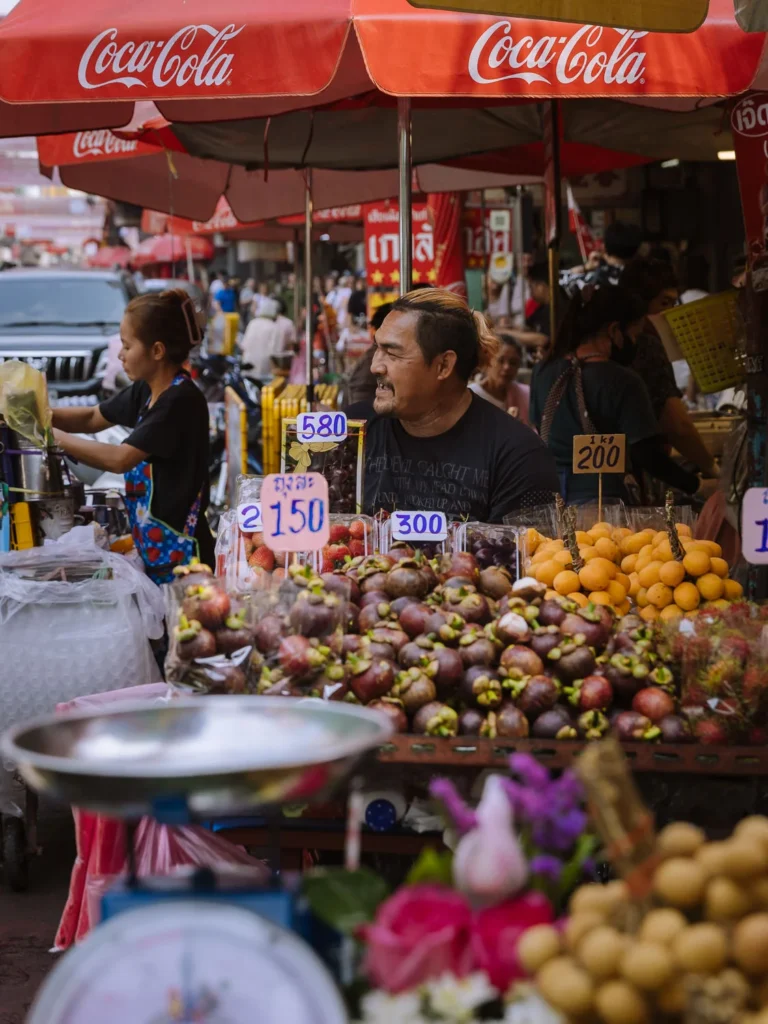 A fruit seller stands behind a stall piled with mangosteens, strawberries, and oranges in Bangkok's Chinatown.