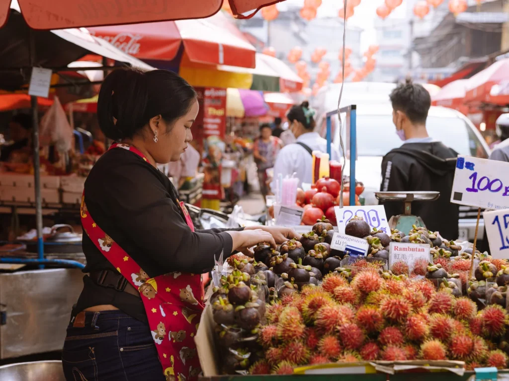 Street vendor arranging mangosteen and rambutan at a busy outdoor market stall with red lanterns and colorful umbrellas overhead.