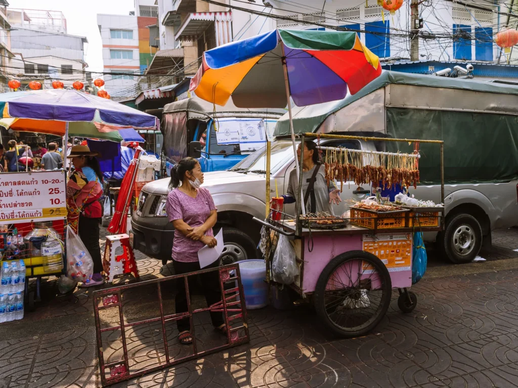 A street food vendor sells grilled meats from a cart beneath a colourful umbrella on a busy Chinatown street, Bangkok.