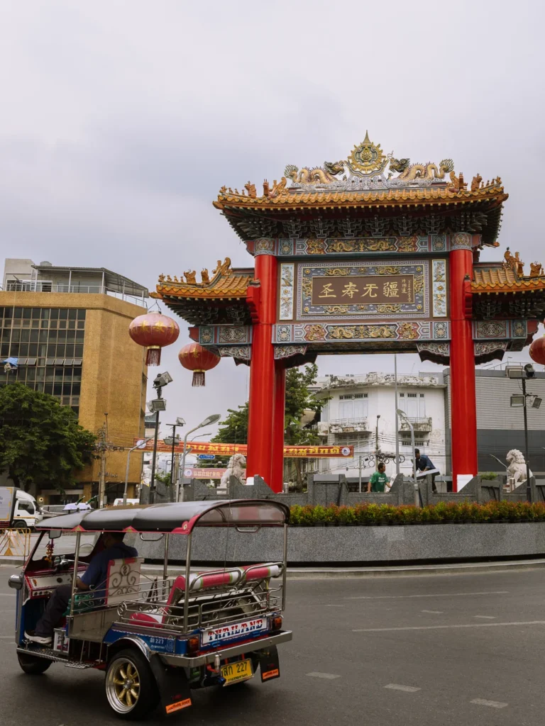 A tuk-tuk passes in front of the ornate red and gold Chinatown Gate on Yaowarat Road, Bangkok, with red lanterns hanging overhead.