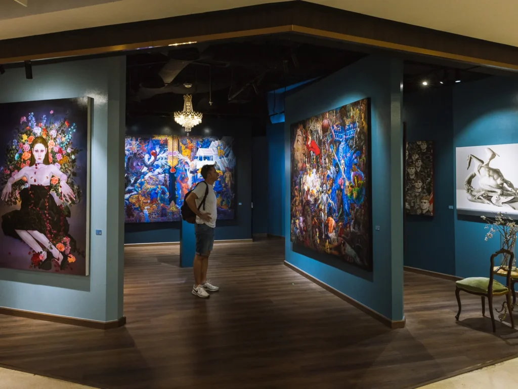 A visitor studies large-scale paintings in a gallery at River City Bangkok, displayed on deep teal walls.