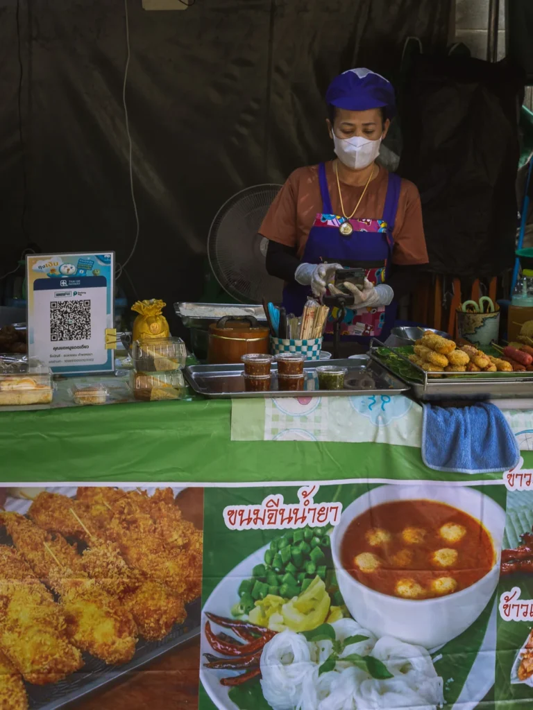 A street food vendor prepares snacks at a stall in Talat Noi, Bangkok, with a menu banner and QR payment sign in the foreground.