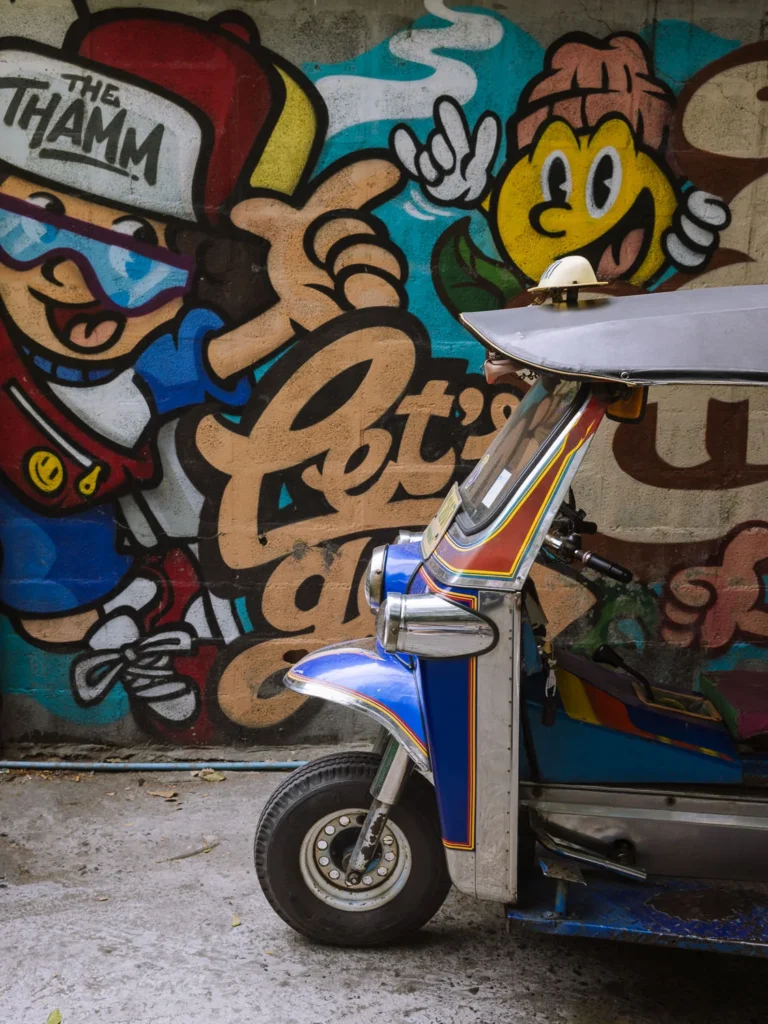 A tuk-tuk parked in front of a bold graffiti mural on a lane wall in Talat Noi, Bangkok.