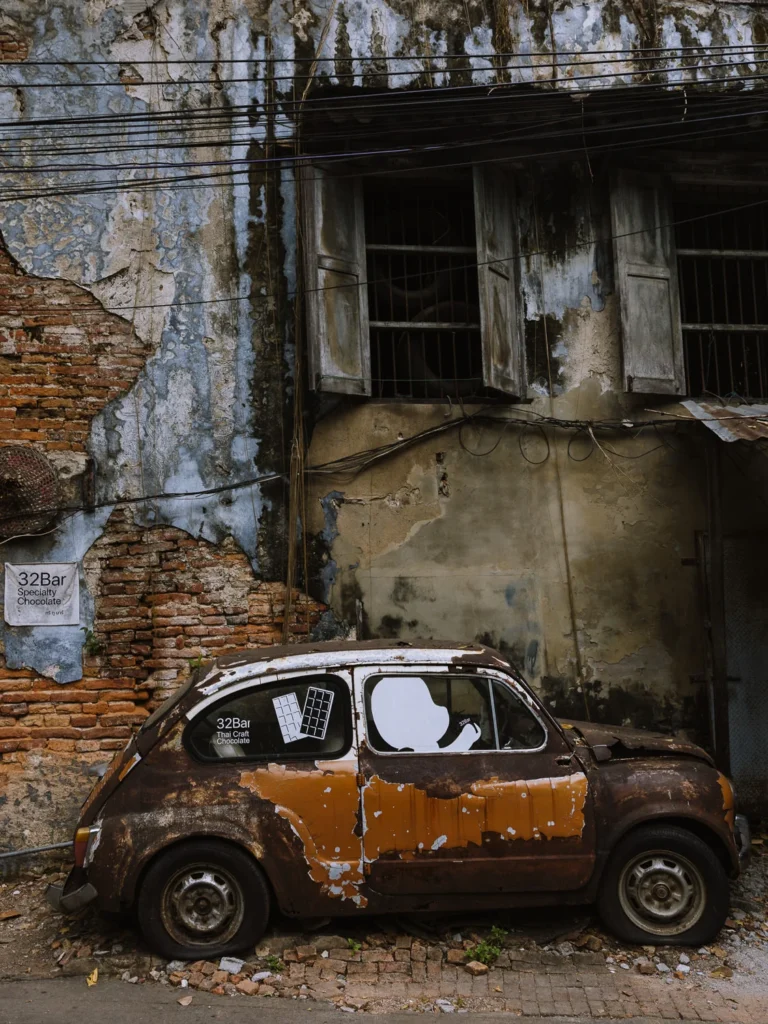 Rusted abandoned car outside a derelict shophouse in Talat Noi, Bangkok, with a 32Bar speciality chocolate sign on the wall.