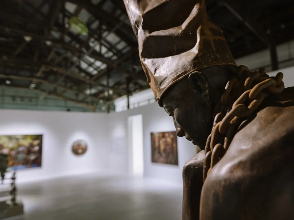 A bronze sculptural figure wearing a chain necklace stands in the foreground of a gallery space at Warehouse 30, Bangkok, with paintings visible on the white walls behind.