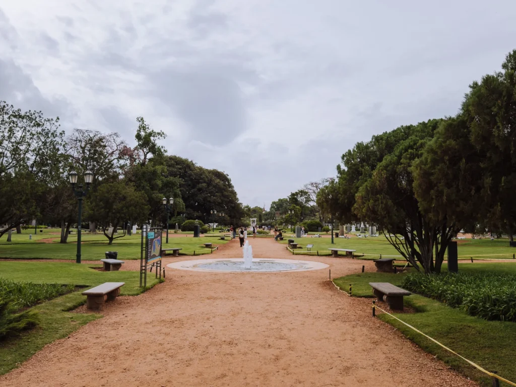 A wide gravel path leading through the Bosques de Palermo in Buenos Aires, with a small circular fountain in the foreground, park benches on either side and visitors strolling in the distance
