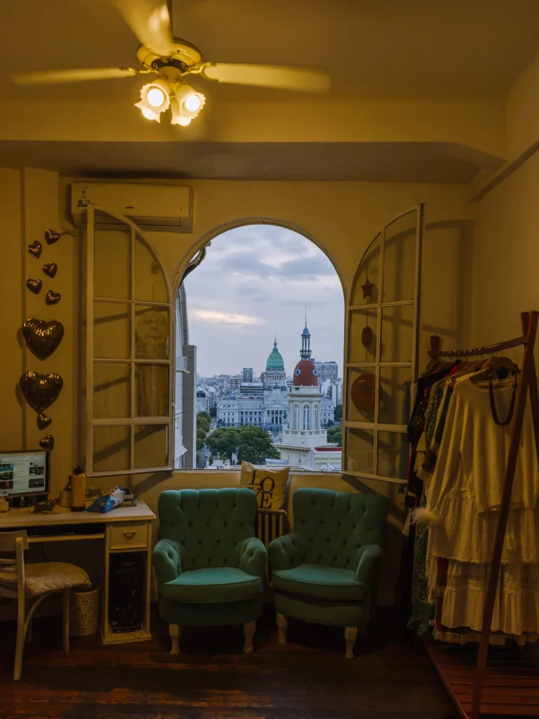 The interior of a Buenos Aires apartment with two teal velvet armchairs facing an arched window, through which the green copper dome and ornate towers of the Palacio del Congreso are visible at dusk, with heart-shaped decorations on the wall and a clothing rail to the right
