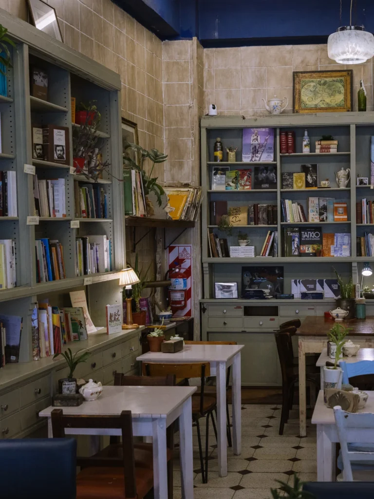 The eclectic interior of a Buenos Aires bookshop-café in San Telmo, with floor-to-ceiling shelves of books, potted plants, mismatched café tables and chairs, and tango books displayed on the shelves