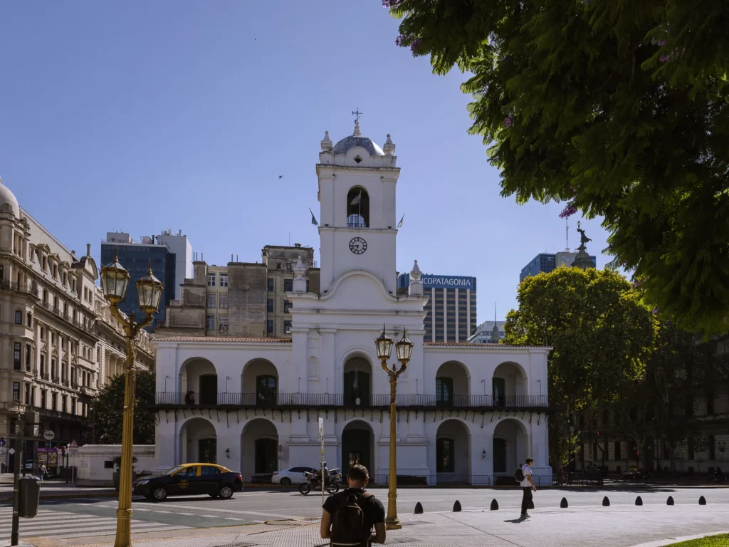 The white colonial façade of the Cabildo on Plaza de Mayo in Buenos Aires, with its arched arcade, central clock tower and gold ornamental lampposts in the foreground against a deep blue sky