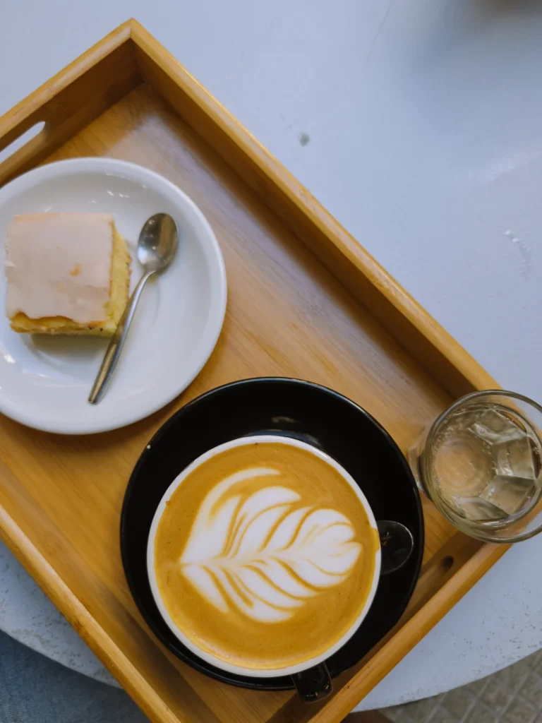 A flat white coffee with leaf latte art served on a wooden tray alongside a slice of cake and a glass of iced water at a speciality café in Buenos Aires