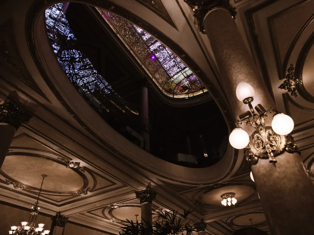 The ornate ceiling of Confitería La Ideal in Buenos Aires, with a large oval stained-glass skylight in blues and purples, gilded plasterwork cornicing and brass wall sconces