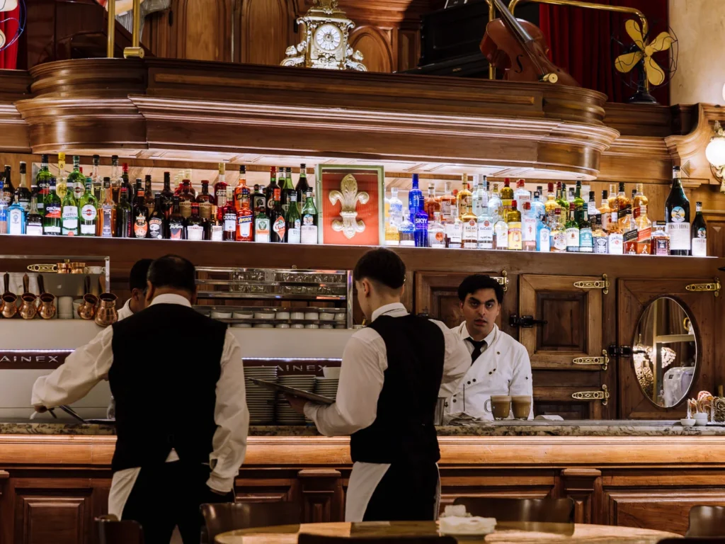 Waiters in black waistcoats working behind the polished wooden bar counter of Confitería La Ideal in Buenos Aires, with a back bar lined with spirits bottles and a vintage clock above