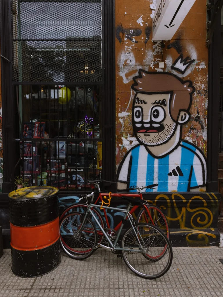 Street art mural of a cartoon figure wearing a blue and white Argentine football shirt with a crown on a weathered building wall in Buenos Aires, with two bicycles locked up in front and a hardware shop window alongside