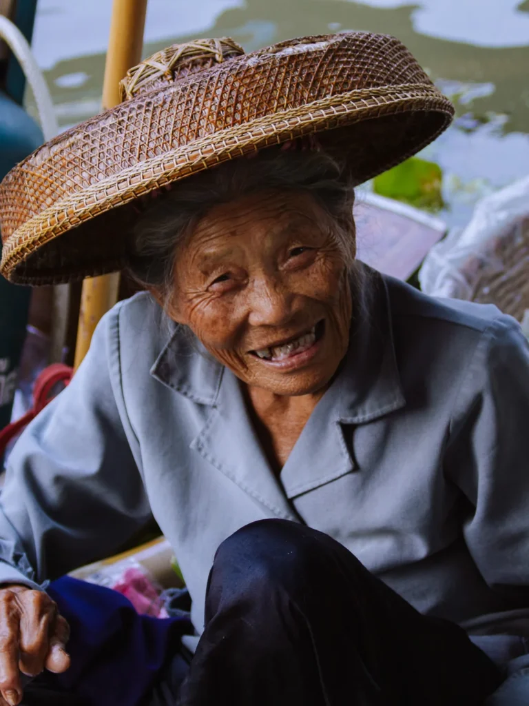 An elderly woman vendor wearing a woven straw hat smiles at a floating market near Bangkok.
