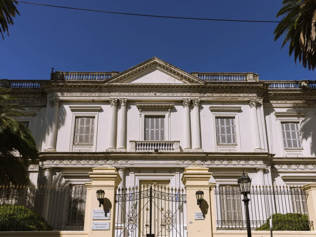 The neoclassical façade of the Servicio Histórico del Ejército (Argentine Army Historical Service) building in Buenos Aires, with Corinthian columns, a triangular pediment, wrought-iron entrance gates and palm trees framing the building against a deep blue sky