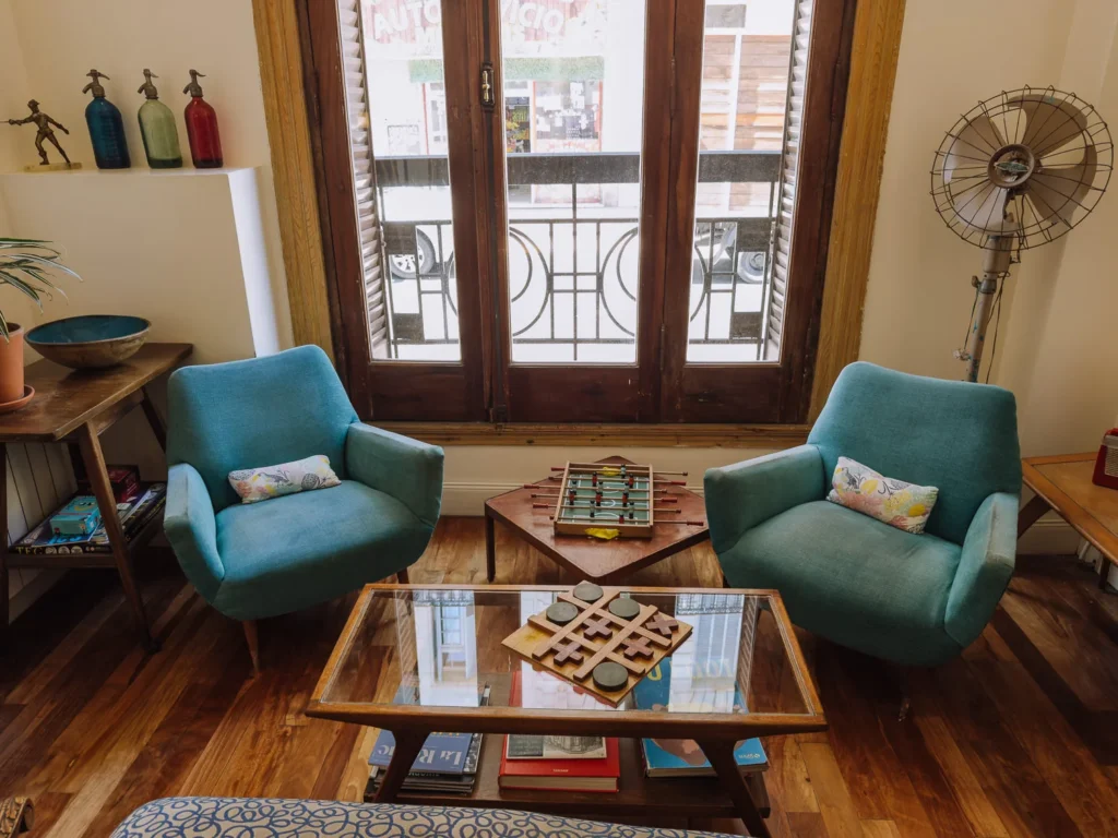 The retro-styled lounge area of a boutique hotel in San Telmo, Buenos Aires, with two teal mid-century armchairs, a glass coffee table with board games, coloured soda siphons on a wooden shelf, and French doors opening onto a wrought-iron balcony