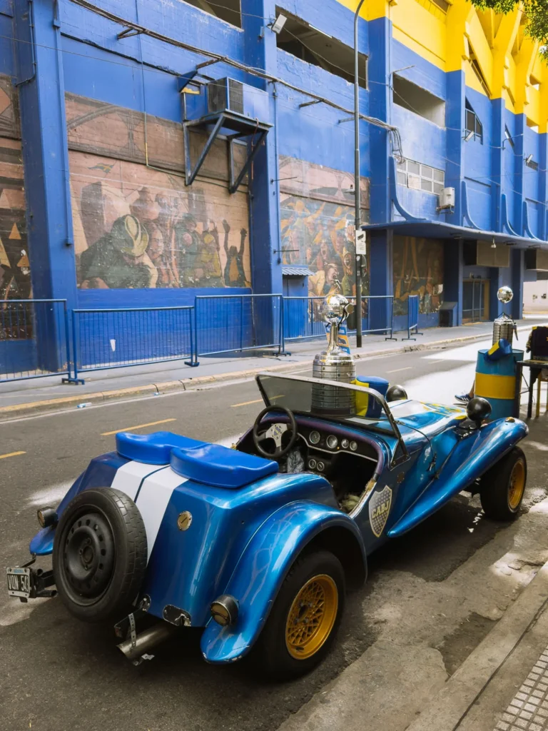 A custom blue and white open-top car painted in Boca Juniors colours parked outside La Bombonera stadium in La Boca, Buenos Aires, with football murals on the stadium wall behind