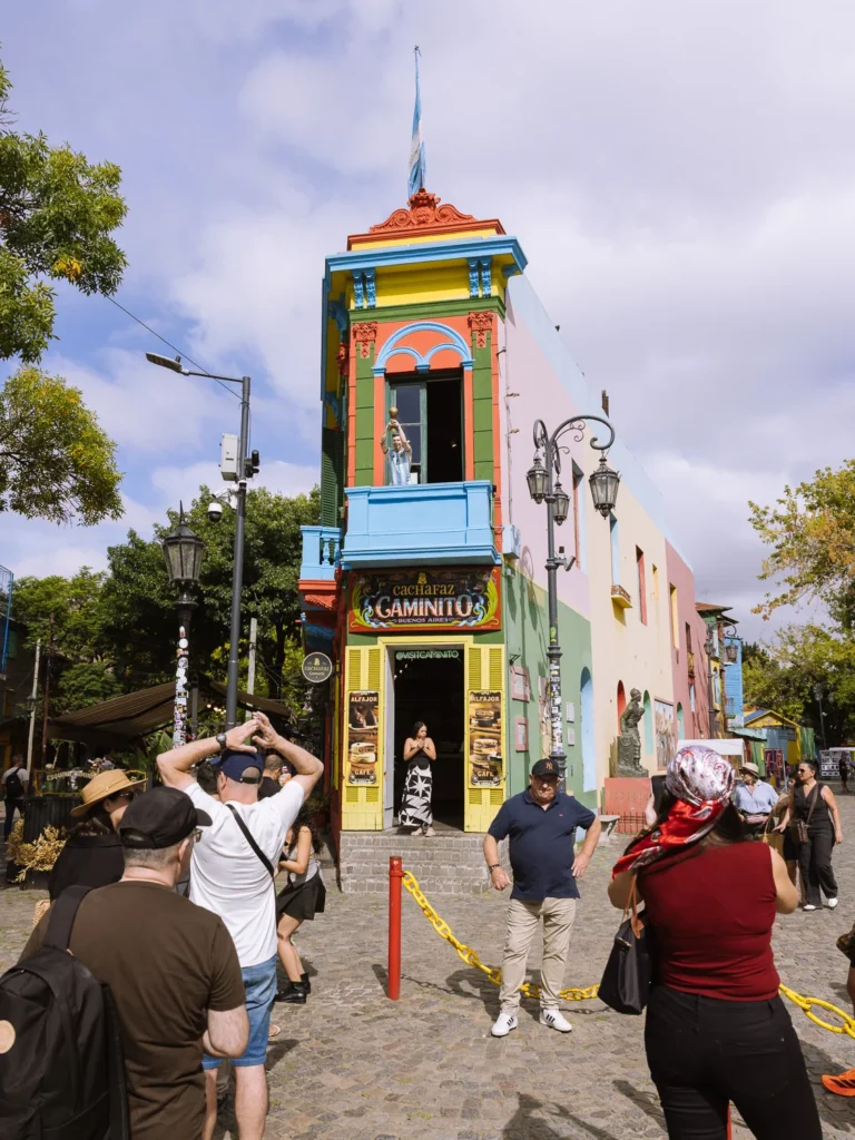 Tourists photographing the iconic multi-coloured corner building at Caminito in La Boca, Buenos Aires, with a Maradona figure in the upper window and a Lionel Messi figure above the Cachafaz café entrance