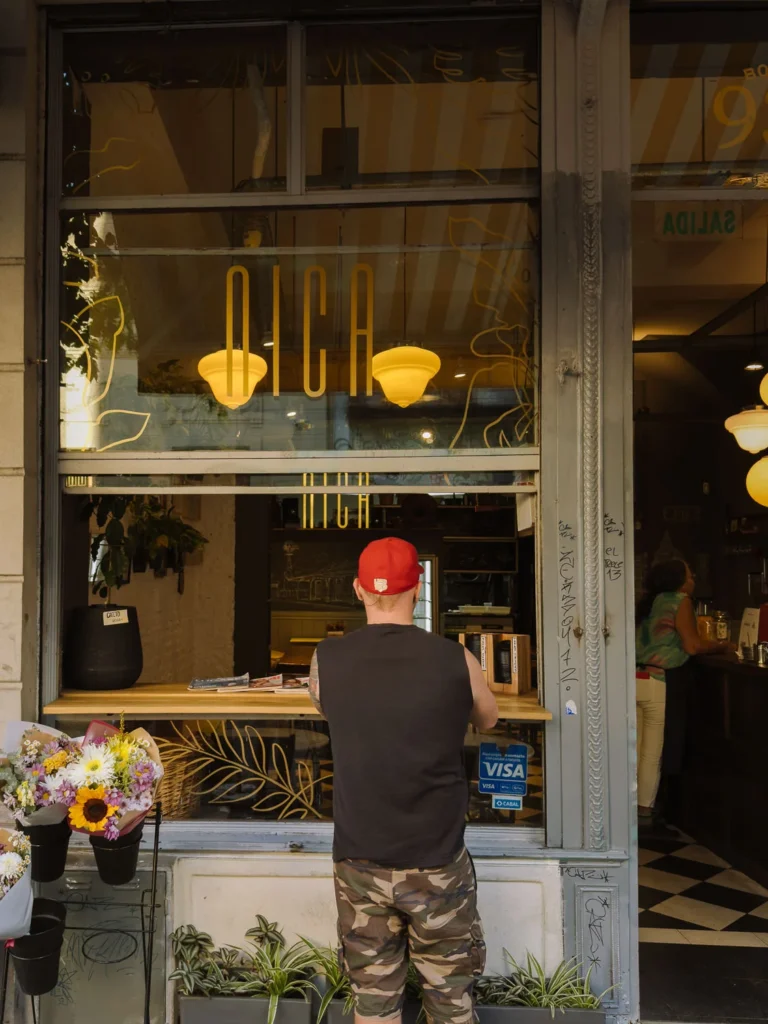 The street-front window of Nica café in Buenos Aires, with a person ordering at the outdoor counter, yellow pendant lights visible inside, and fresh flowers displayed beside the entrance