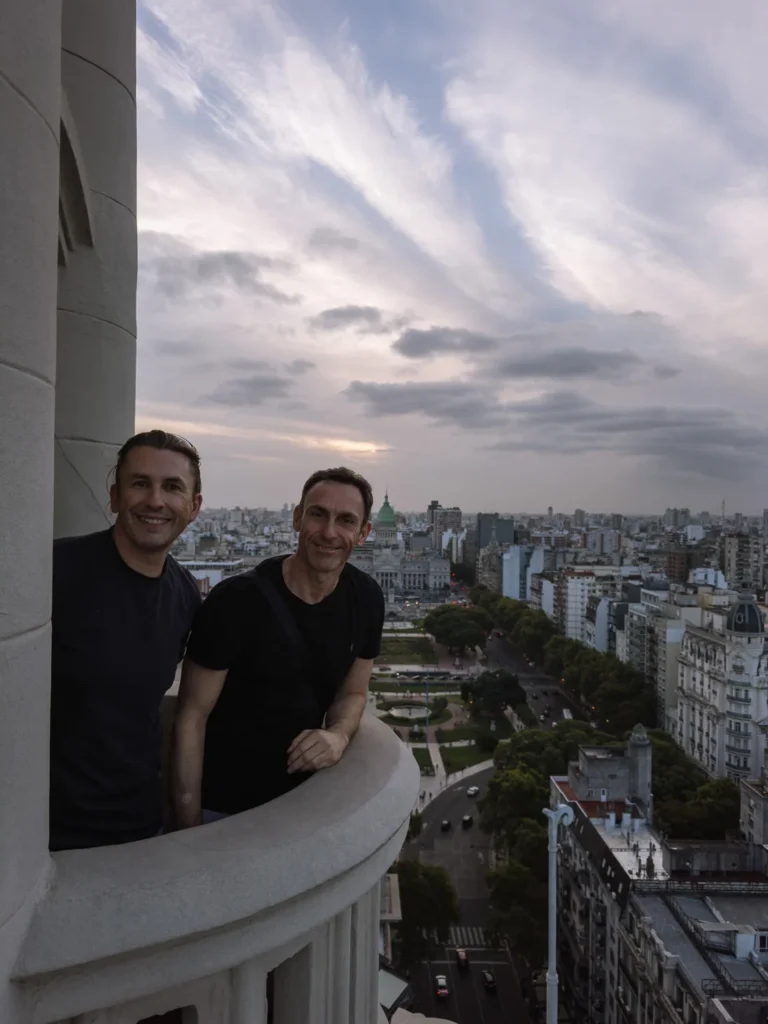 Paul and Mark on the lighthouse balcony of Palacio Barolo in Buenos Aires, smiling with the city skyline and the dome of the National Congress visible behind them at dusk