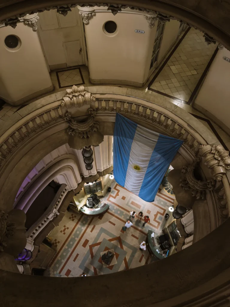 Looking down through the ornate stairwell atrium of Palacio Barolo in Buenos Aires, with a large Argentine flag hanging in the centre above the decorated geometric floor below