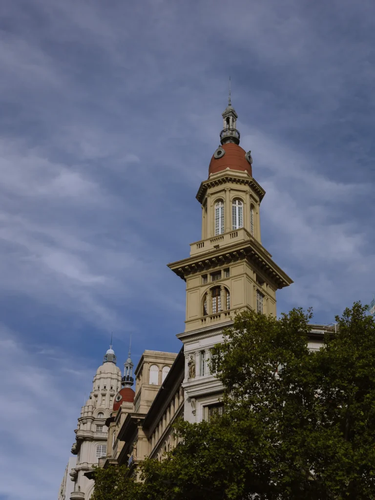 The ornate tower of the Palacio Barolo on Avenida de Mayo, Buenos Aires, with its terracotta copper dome and Beaux-Arts stonework rising above the tree canopy against a dramatic blue sky with wispy clouds