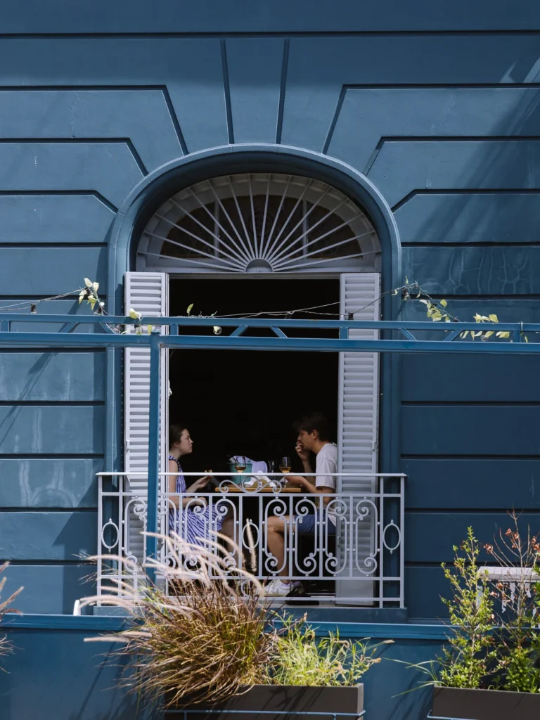 A couple dining at a table on a wrought-iron balcony of a blue Beaux-Arts building in Palermo, Buenos Aires, with arched shuttered doors and plants below