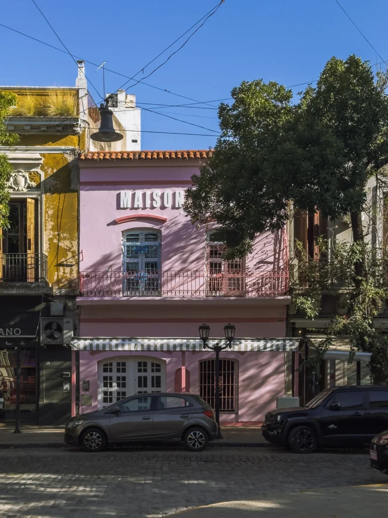 The pink façade of Maison restaurant on a cobblestone street in San Telmo, Buenos Aires, with a striped awning, wrought-iron balcony railings and tree shadows across the frontage on a sunny day