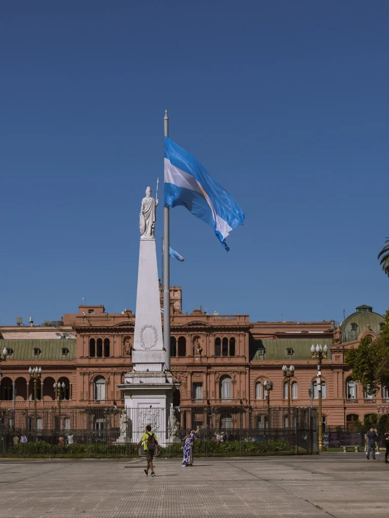 The Pirámide de Mayo monument in Plaza de Mayo, Buenos Aires, with the Argentine flag flying from a tall flagpole and the pink facade of the Casa Rosada visible in the background