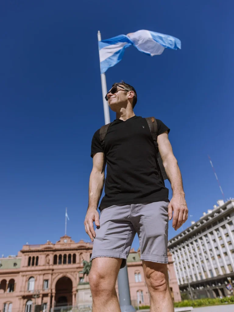 Mark standing in Plaza de Mayo, Buenos Aires, with the Argentine flag and the Casa Rosada presidential palace visible in the background against a deep blue sky