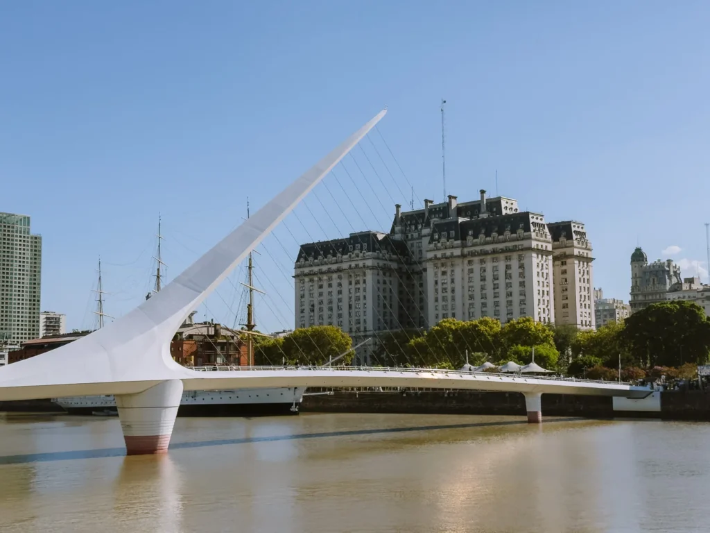The Puente de la Mujer suspension bridge in Puerto Madero, Buenos Aires, with its distinctive white angled pylon and steel cables, with a tall ship moored behind and the Edificio Libertador visible across the water against a clear blue sky
