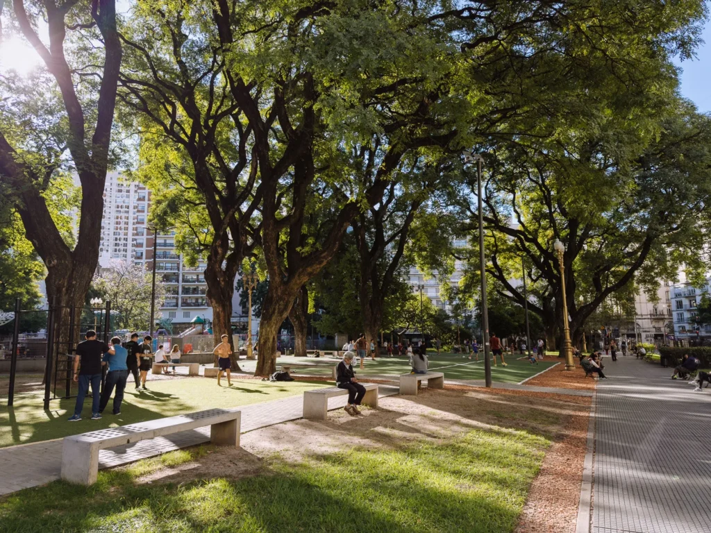 Locals relaxing on benches and lawns beneath large shade trees in a sunlit Buenos Aires public plaza, with apartment buildings and ornate lampposts visible through the canopy