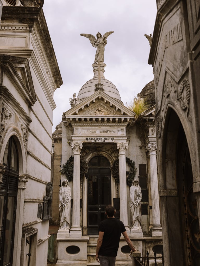 Paul standing before the grand mausoleum of Bernardo de Irigoyen at Recoleta Cemetery, Buenos Aires, flanked by marble angel statues and topped with a winged figure