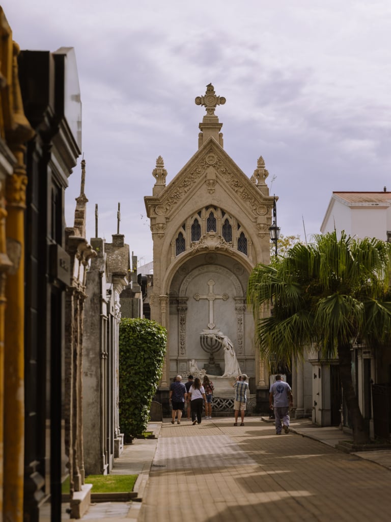 Visitors walking through the narrow streets of Recoleta Cemetery in Buenos Aires, towards an elaborate Gothic-style marble mausoleum with a carved cross and arched facade