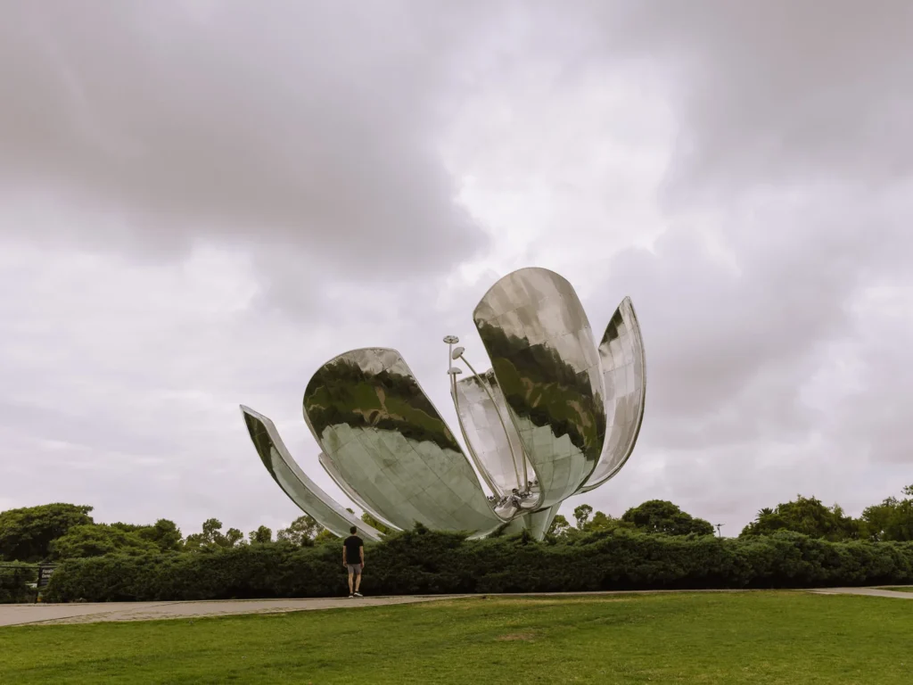 Mark standing beside the Floralis Genérica sculpture in Recoleta, Buenos Aires — a giant polished steel flower with open petals reflecting the overcast sky, surrounded by green lawns